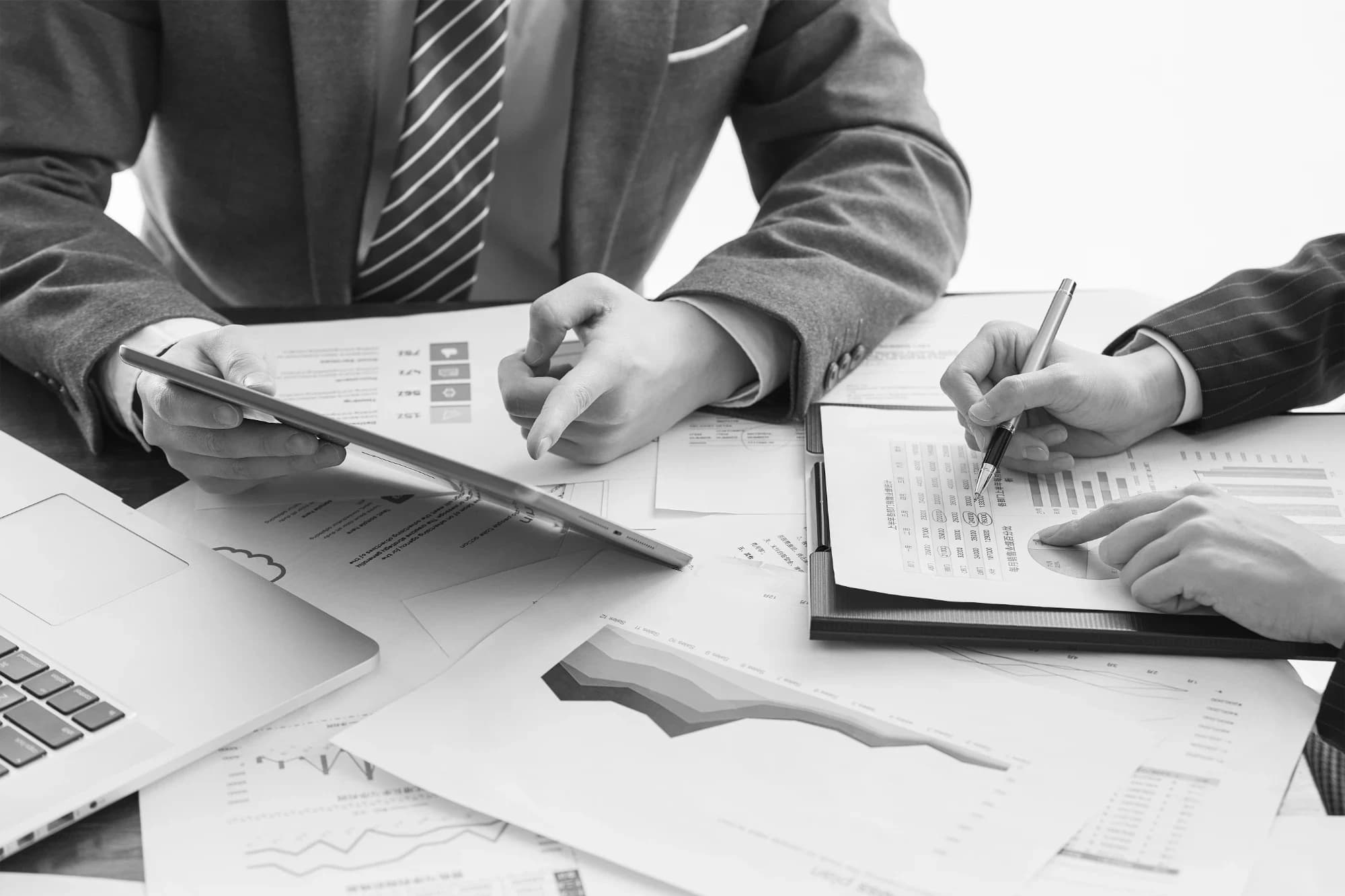 Colleagues in business attire review printed financial charts and reports with a tablet and laptop